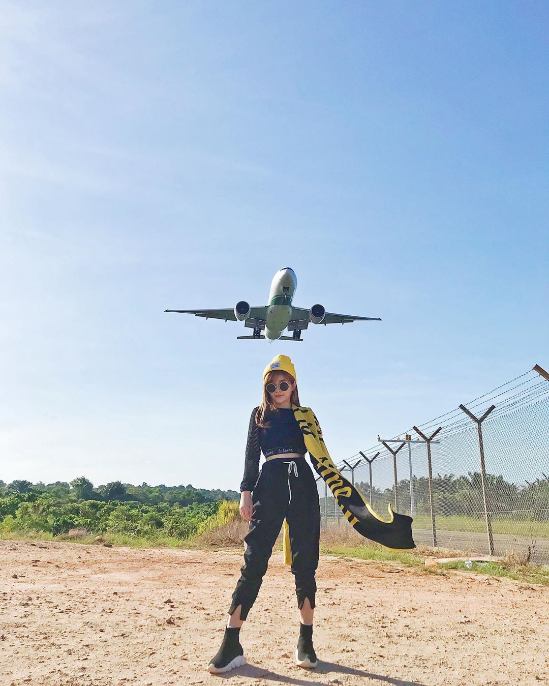 A woman standing for a photograph as an airplane flies overhead. 